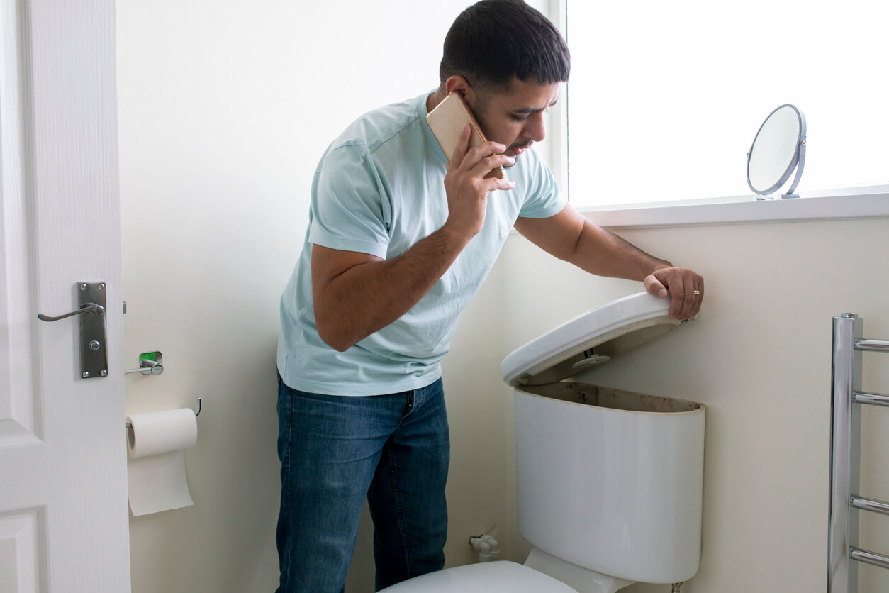 A man talks on the phone while looking into the cistern tank of a faulty toilet.